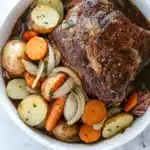 3 packet pot roast slow cooker - the image shows a large white bowl filled with a dish of beef stew. the beef appears to be cooked medium-rare and has a rich, dark brown color. it is garnished with sliced onions, carrots, and potatoes. the dish is served on a white marble countertop with a blue and white striped napkin on the side. there are also some sprigs of parsley scattered around the bowl. the overall appearance of the dish is hearty and appetizing.
