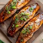 Air fryer salmon teriyaki - the image shows three pieces of salmon on a wooden cutting board. the salmon is cooked to a golden brown color and is covered in a sesame seeds and green onions. the cutting board is placed on a light-colored wooden table with a few sprigs of dill scattered around it. the background is blurred, but it appears to be a kitchen countertop. the overall aesthetic of the image is rustic and appetizing.