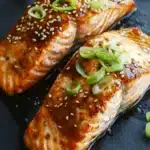 Air fryer teriyaki salmon - the image shows two pieces of salmon on a black slate plate. the salmon is cooked to a golden brown color and is covered in sesame seeds and green onions. the sauce is drizzled over the salmon, giving it a glossy sheen. there are also a few small pieces of sliced green onions scattered around the plate, adding a pop of color to the dish. the background is blurred, making the salmon the focal point of the image.