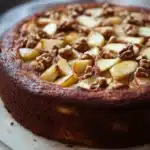 Apple cake yogurt - the image is of a round cake on a wooden cutting board. the cake appears to be a chocolate cake with a golden brown crust. on top of the cake, there are sliced apples arranged in a circular pattern, with some of them overlapping each other. the apples are cut into small cubes and are scattered throughout the cake. there are also some chopped nuts on top, adding a crunchy texture to the dish. in the background, there is a white bowl and a white cloth napkin, suggesting that the cake is being prepared for baking.