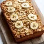 Banana bread baked oatmeal - the image shows a rectangular loaf of bread on a wooden cutting board. the bread appears to be freshly baked and has a golden brown crust on top. on top of the bread, there are several sliced bananas arranged in a circular pattern. the bananas are cut into thin slices and are placed on top of a layer of oats. the oats are scattered throughout the bread and are a light brown color. the cutting board is placed on a white marble countertop with a white towel on the side.