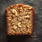 Banana cake slice plating - the image is a close-up of a slice of bread on a black textured surface. the bread appears to be freshly baked and has a golden brown color. it is topped with chopped walnuts, giving it a crunchy texture. the walnuts are arranged in a scattered manner, with some overlapping each other. the edges of the bread are slightly curled, indicating that it has been freshly baked. the background is blurred, making the bread the focal point of the image.
