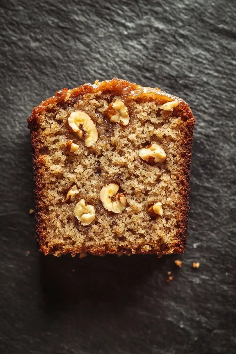Banana cake slice plating - the image is a close-up of a slice of bread on a black textured surface. the bread appears to be freshly baked and has a golden brown color. it is topped with chopped walnuts, giving it a crunchy texture. the walnuts are arranged in a scattered manner, with some overlapping each other. the edges of the bread are slightly curled, indicating that it has been freshly baked. the background is blurred, making the bread the focal point of the image.