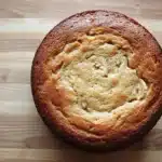 Banana cake toddler - the image is a close-up of a round cake on a wooden surface. the cake appears to be freshly baked and has a golden brown crust on top. the center of the cake is filled with a creamy, light-colored filling, which looks like banana slices. the filling is spread evenly across the cake, creating a swirl pattern. the background is blurred, making the cake the focal point of the image.