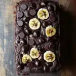 Banana chocolate cake - the image is a close-up of a rectangular piece of chocolate cake on a wooden surface. the cake appears to be freshly baked and has a rich, dark brown color. on top of the cake, there are several sliced bananas arranged in a circular pattern. the bananas are cut into small pieces and are scattered around the cake. there are also a few chocolate chips scattered around them. the chocolate chips are dark brown and appear to be coated in chocolate. the background is blurred, making the cake the focal point of the image.