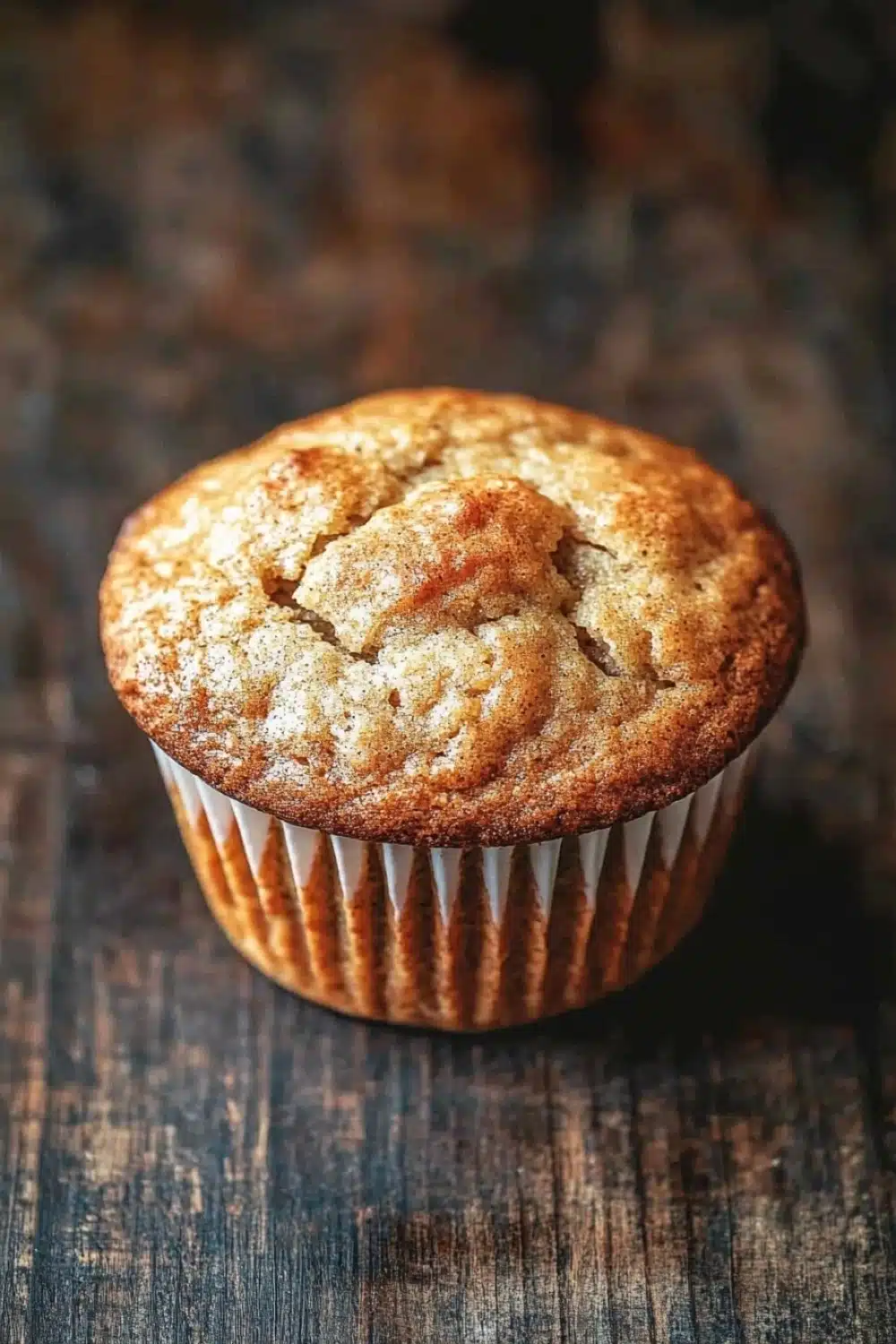 Banana muffin gluten free - the image is a close-up of a freshly baked muffin on a wooden surface. the muffin is round and golden brown in color, with a crumbly texture on top. it appears to be freshly baked and has a soft, fluffy interior. the edges of the muffin are lined with white paper liners. the background is blurred, but it seems to be a rustic, weathered wooden table. the overall mood of the image is warm and inviting.