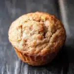 Banana muffin no added sugar - the image is a close-up of a freshly baked muffin on a wooden surface. the muffin appears to be golden brown in color and has a crumbly texture. it is sitting on a dark wooden surface with a rustic, weathered appearance. the background is blurred, making the muffin the focal point of the image.