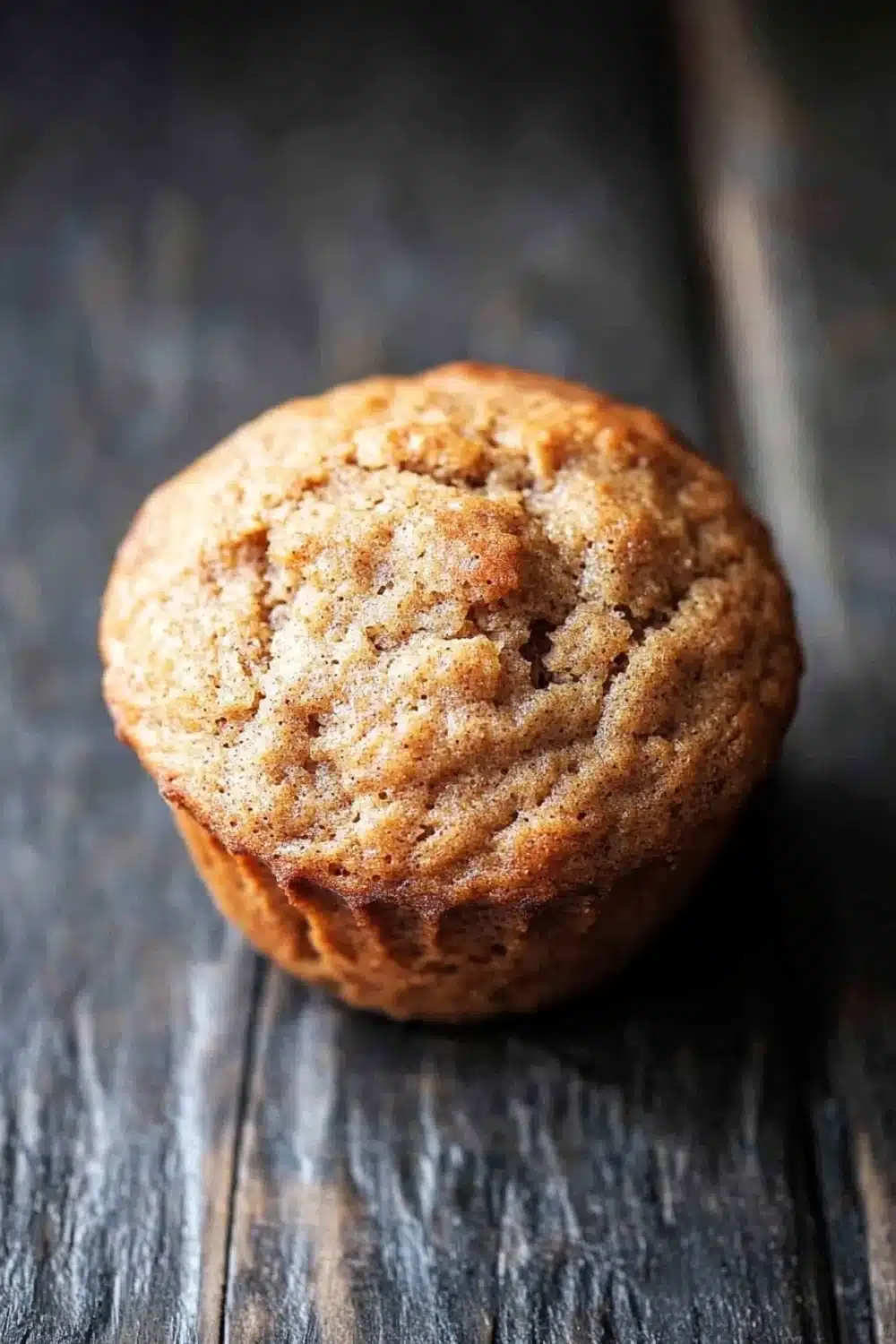 Banana muffin no added sugar - the image is a close-up of a freshly baked muffin on a wooden surface. the muffin appears to be golden brown in color and has a crumbly texture. it is sitting on a dark wooden surface with a rustic, weathered appearance. the background is blurred, making the muffin the focal point of the image.
