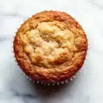 Banana muffin protein - the image is a close-up of a muffin on a white marble surface. the muffin appears to be freshly baked and has a golden brown color. it has a crumbly texture on top, with a light dusting of powdered sugar on top. the edges of the muffin are slightly curled, indicating that it has been freshly baked. the background is blurred, making the muffins the focal point of the image.