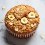 Banana oat yogurt muffin - the image is of a freshly baked muffin on a white marble surface. the muffin is golden brown in color and has a crumbly texture. on top of the muffin, there are three sliced bananas arranged in a circular pattern. the bananas are cut in half and are placed on top of a pile of oats. the oats are scattered around the muffins, giving the image a rustic and natural feel.