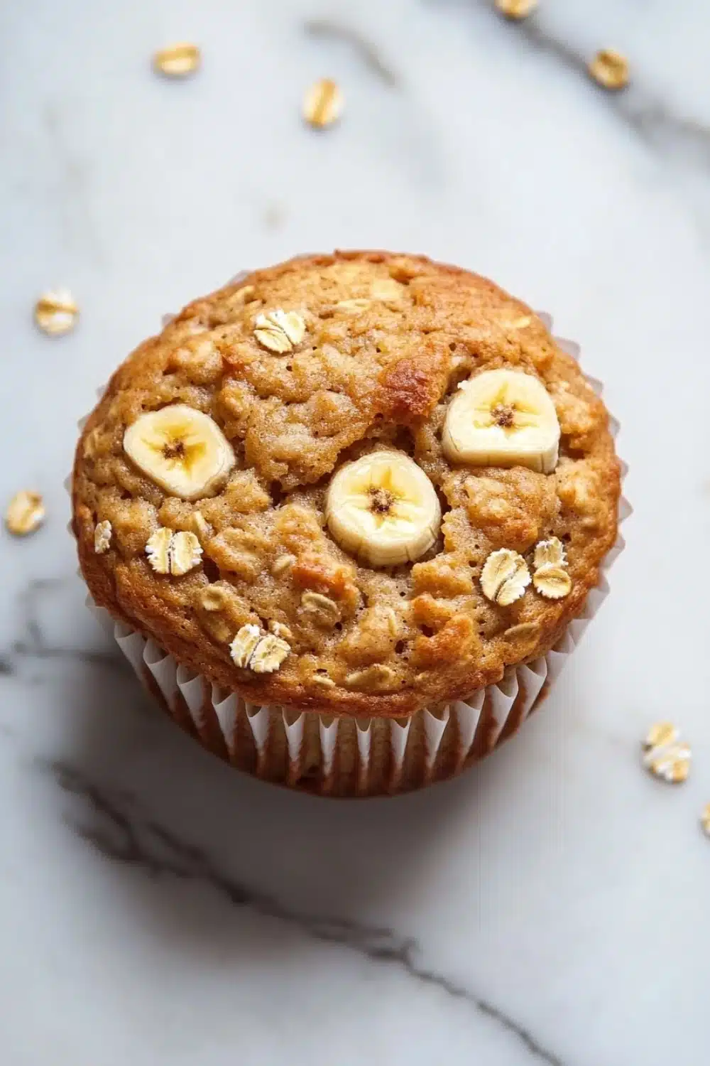 Banana oat yogurt muffin - the image is of a freshly baked muffin on a white marble surface. the muffin is golden brown in color and has a crumbly texture. on top of the muffin, there are three sliced bananas arranged in a circular pattern. the bananas are cut in half and are placed on top of a pile of oats. the oats are scattered around the muffins, giving the image a rustic and natural feel.