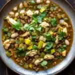 Beef green chicken chili freezer meal - the image is a close-up of a bowl of soup on a wooden table. the soup appears to be a stew or stew with chunks of chicken, beans, and vegetables mixed in. the bowl is made of dark brown ceramic and is garnished with fresh cilantro. the vegetables include white beans, yellow bell peppers, and green onions. the broth is thick and thick, and the soup looks creamy and inviting. there are a few sprigs of cilantro scattered around the bowl, adding a pop of green color to the dish.