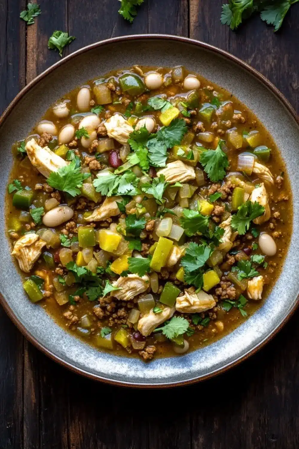 Beef green chicken chili freezer meal - the image is a close-up of a bowl of soup on a wooden table. the soup appears to be a stew or stew with chunks of chicken, beans, and vegetables mixed in. the bowl is made of dark brown ceramic and is garnished with fresh cilantro. the vegetables include white beans, yellow bell peppers, and green onions. the broth is thick and thick, and the soup looks creamy and inviting. there are a few sprigs of cilantro scattered around the bowl, adding a pop of green color to the dish.