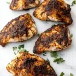 Blackened tilapia in air fryer - the image shows four pieces of grilled chicken breasts on a white marble surface. the chicken breasts are golden brown and appear to be seasoned with herbs and spices. there are also a few sprigs of parsley scattered around the surface, adding a pop of green color to the dish. the background is blurred, making the chicken breasts the focal point of the image.