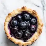 Blueberry muffin mix cobbler - the image is a close-up of a blueberry tart on a white marble surface. the tart has a golden-brown crust and is filled with fresh blueberries. the blueberries are arranged in a circular pattern around the edges of the tart, with some of them overlapping each other. the filling is a deep purple color, and the tart appears to be freshly baked. the background is blurred, making the tart the focal point of the image.
