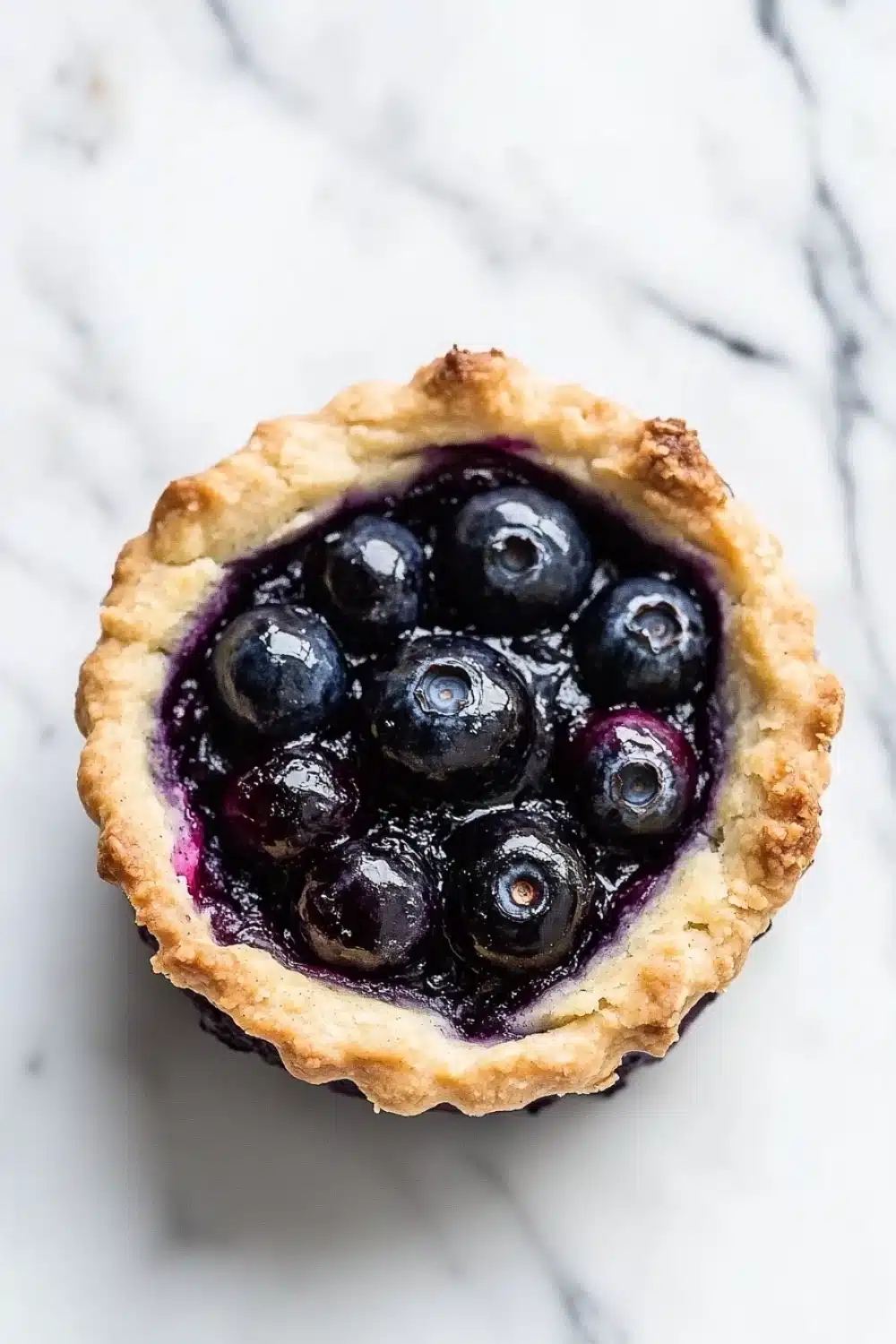 Blueberry muffin mix cobbler - the image is a close-up of a blueberry tart on a white marble surface. the tart has a golden-brown crust and is filled with fresh blueberries. the blueberries are arranged in a circular pattern around the edges of the tart, with some of them overlapping each other. the filling is a deep purple color, and the tart appears to be freshly baked. the background is blurred, making the tart the focal point of the image.