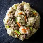 Chicken and beef dumplings crockpot freezer meal - the image is a close-up of a dish on a black slate plate. the dish appears to be a type of ravioli, with a creamy white sauce on top. the sauce is drizzled over the top of the raviolis and is garnished with chopped green onions, carrots, and chunks of meat. the meat is cooked to a medium-rare and looks tender and juicy. the plate is round and has a smooth texture.