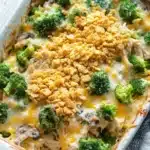 Chicken and broccoli casserole - the image shows a white rectangular baking dish filled with a casserole dish. the dish appears to be made with broccoli, chicken, and cheese. the broccoli is cut into small pieces and is scattered throughout the dish, with some pieces of chicken and mushrooms visible. the cheese is melted and bubbly, and there is a golden brown crumbly topping on top. the baking dish is sitting on a wooden table with a gray and white striped napkin next to it.