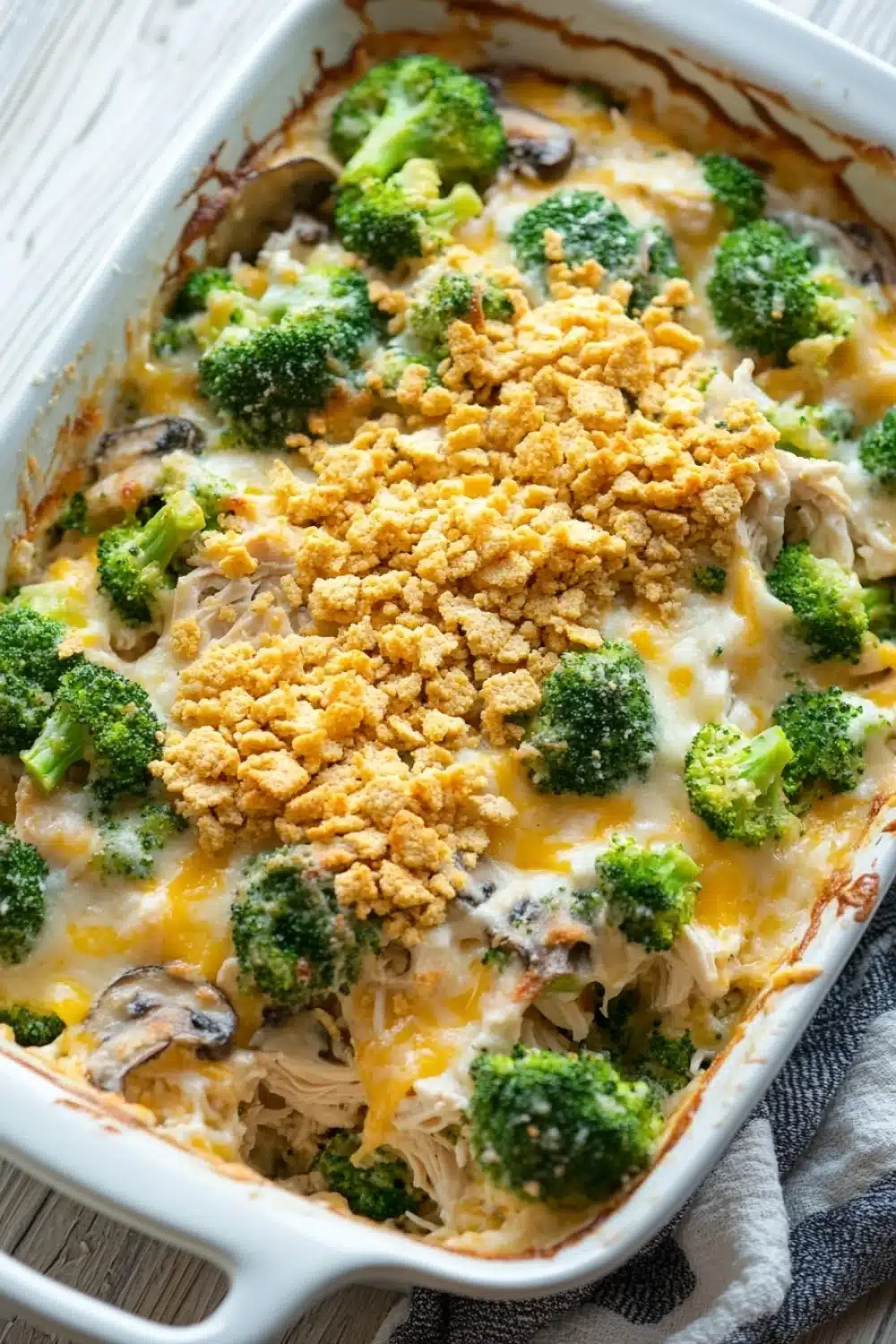 Chicken and broccoli casserole - the image shows a white rectangular baking dish filled with a casserole dish. the dish appears to be made with broccoli, chicken, and cheese. the broccoli is cut into small pieces and is scattered throughout the dish, with some pieces of chicken and mushrooms visible. the cheese is melted and bubbly, and there is a golden brown crumbly topping on top. the baking dish is sitting on a wooden table with a gray and white striped napkin next to it.