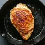 Chicken breast in cast iron skillet - the image shows a piece of cooked chicken in a black cast iron skillet. the chicken appears to be seasoned with herbs and spices, giving it a golden brown color. the skillet is sitting on a dark grey countertop, and the chicken is glistening with oil. the overall appearance of the dish is savory and appetizing.