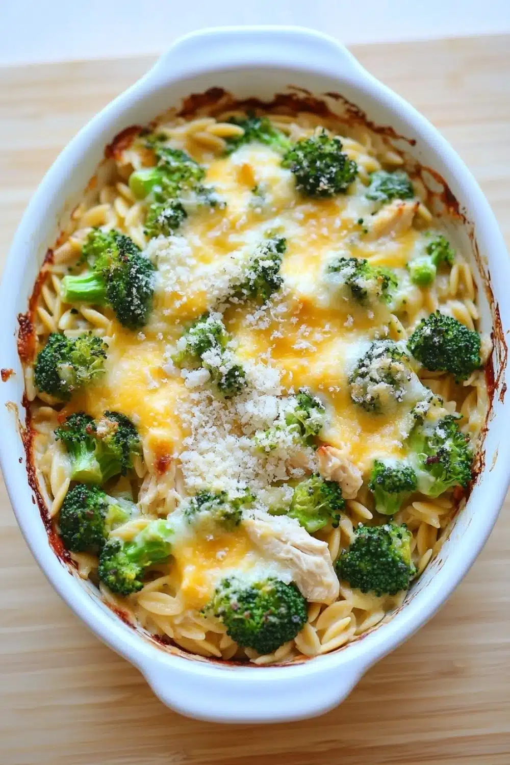 Chicken broccoli casserole with orzo - the image shows a white ceramic casserole dish filled with a dish of pasta and broccoli. the pasta appears to be fusilli, which is a type of spiral-shaped pasta, and it is topped with melted cheese and grated parmesan cheese. the broccoli is bright green and looks fresh and healthy. the dish is sitting on a wooden cutting board.