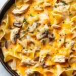 Chicken noodle casserole skillet - the image shows a skillet filled with a creamy chicken noodle casserole. the dish is made up of fettuccine noodles, chunks of chicken, and chunks of mushrooms. the noodles are cooked al dente and are covered in a yellow sauce with melted cheese and herbs sprinkled on top. the skillet is black and has a handle on one side. the background is a white marble countertop.