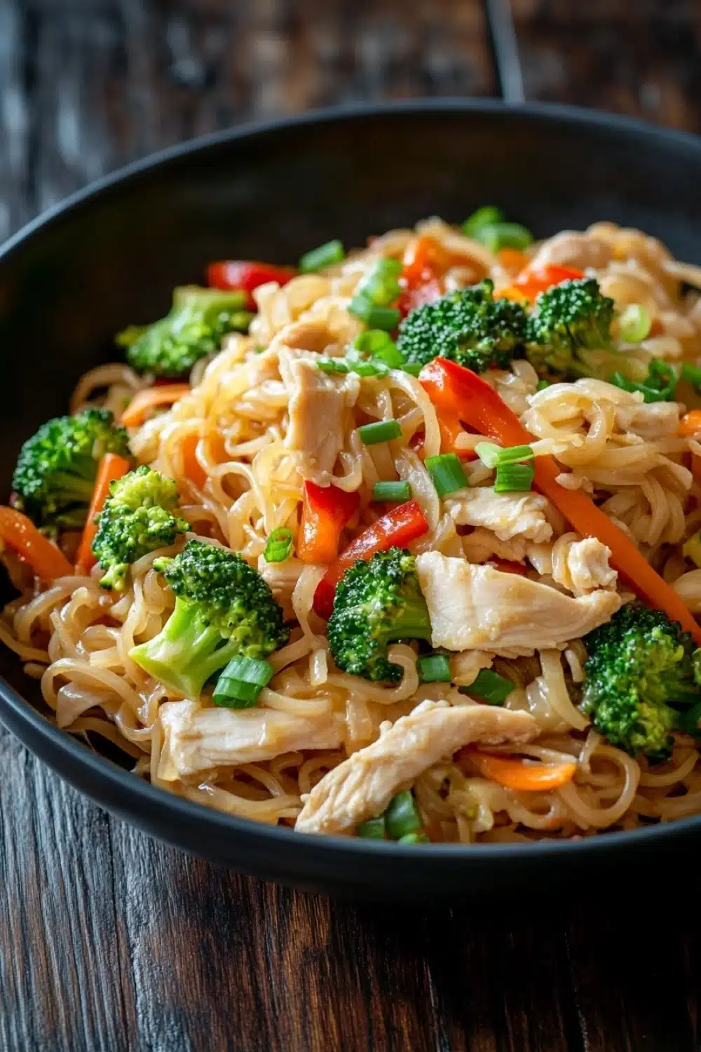 Chicken noodles low calorie - the image is a close-up of a bowl of noodles with chicken and vegetables. the noodles are light brown and appear to be stir-fry. the chicken is cut into small pieces and is mixed with various vegetables such as broccoli, carrots, and bell peppers. the vegetables are bright green and look fresh and vibrant. the dish is served in a black bowl with a wooden table in the background.