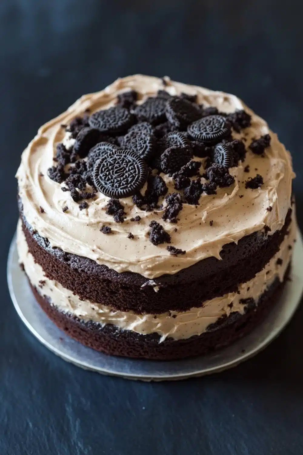 Chocolate cake oreo frosting - the image is of a round chocolate cake on a silver cake stand. the cake is covered in a light brown frosting with a swirl of cream cheese frosting on top. on top of the frosting, there are several oreo cookies arranged in a circular pattern. the cookies are dark brown in color and appear to be freshly baked. the background is a dark grey countertop.
