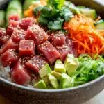 Classic tuna poke bowl - the image is a close-up of a bowl of food. the bowl is made of dark brown ceramic and is placed on a wooden table. the food in the bowl appears to be a salad with a variety of ingredients. on top of the salad, there are sliced tuna, sliced cucumbers, carrots, and avocado. the tuna is covered in sesame seeds and is arranged in a circular pattern. there are also some green herbs scattered throughout the salad. the colors of the ingredients are vibrant and the dish looks fresh and appetizing.