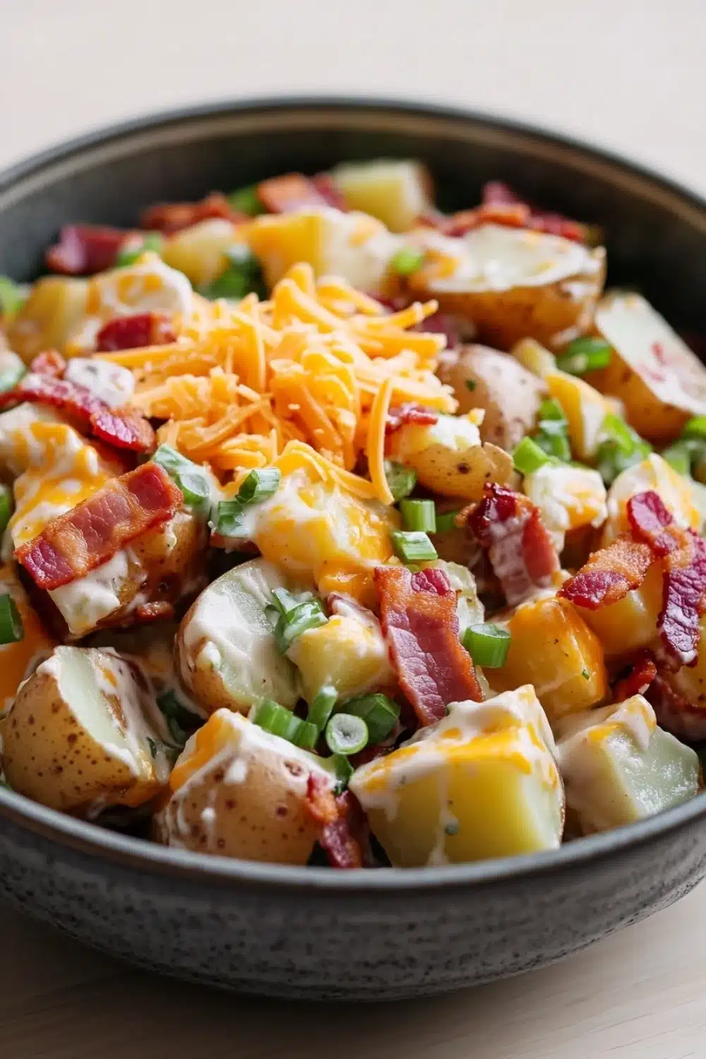 Creamy loaded baked potato salad - the image is a close-up of a bowl of potato salad. the salad is made up of diced potatoes, bacon, and shredded cheese. the potatoes are golden brown and appear to be seasoned with herbs and spices. the bacon is crispy and adds a pop of color to the dish. the cheese is melted and bubbly, and it is drizzled over the top. the bowl is black and has a speckled texture. the background is a wooden table.