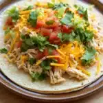 Crock pot chicken tacos - the image shows a plate of food on a wooden table. the plate is round and has a brown rim. on top of the plate, there is a large tortilla filled with shredded chicken, diced tomatoes, and cilantro. the chicken appears to be cooked and is mixed with diced tomatoes and shredded cheese. the cilantro is scattered on top, adding a pop of green color to the dish. the overall appearance of the dish is fresh and appetizing.