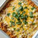 Dorito casserole freezer meal - the image shows a white rectangular baking dish filled with a casserole dish. the dish appears to be made with ground beef, cheese, and other ingredients. the top of the dish is covered in a layer of melted cheese, which is a bright yellow color. there are also a few sprigs of fresh cilantro scattered on top, adding a pop of green color to the dish. on the side, there is a bed of orange-colored ground meat, which looks like ground beef. the casserolle dish is sitting on a wooden table.