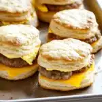Freezer breakfast sandwiches biscuits - the image shows a baking tray with six biscuits on it. the biscuits are golden brown and appear to be freshly baked. they are arranged in a single layer on the tray, with the top biscuit slightly overlapping the bottom one. each biscuit has a bite taken out of it, revealing a generous amount of melted cheese on top. the cheese is a bright yellow color and is oozing out of the sides of the biscuits. the tray is sitting on a white countertop.