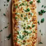 Garlic bread with pizza dough - the image shows a long, rectangular loaf of bread on a wooden cutting board. the bread appears to be freshly baked and has a golden brown crust on top. it is topped with melted cheese and fresh herbs, including chopped parsley. the cheese is melted and bubbly, and there are small pieces of bread scattered around the bread. the cutting board has a light-colored wood grain pattern, and the background is blurred, making the bread the focal point of the image.