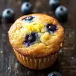 Gluten free lemon blueberry muffin - the image is of a freshly baked blueberry muffin on a wooden surface. the muffin is golden brown and has a crumbly texture. it is topped with fresh blueberries and a sprinkle of yellow zest. there are a few whole blueberries scattered around the muffin, adding a pop of color to the image. the background is blurred, but it appears to be a rustic wooden table.