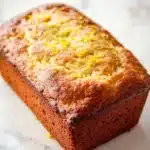 Greek yogurt lemon loaf cake - the image is a close-up of a freshly baked loaf of bread on a white marble countertop. the bread appears to be golden brown in color and has a slightly crumbly texture. on top of the bread, there is a layer of yellow zest, which adds a pop of color to the loaf. the zest is scattered across the top, creating a textured pattern. the loaf is slightly tilted to the side, and the edges are slightly curled. the background is blurred, making the bread the focal point of the image.