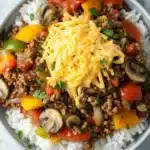 Ground beef dinner make ahead - the image is a close-up of a bowl of food. the bowl is made of stainless steel and is sitting on a white marble countertop. the food in the bowl appears to be a dish made with ground beef, diced tomatoes, mushrooms, and other vegetables. there is a layer of shredded cheese on top of the dish. the colors of the ingredients are vibrant and the dish looks appetizing.