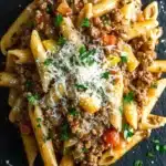 Ground beef pasta dish - the image shows a plate of penne pasta with ground beef and tomato sauce. the pasta is cooked al dente and is arranged in a circular pattern on a black slate plate. the dish is garnished with chopped parsley and grated parmesan cheese. the ground beef is a rich, dark brown color and appears to be cooked until it is thick and tender. the tomato sauce is a bright red color and is scattered throughout the dish. the overall appearance of the dish is hearty and appetizing.