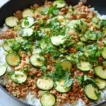 Ground chicken zucchini rice skillet - the image shows a black frying pan with a serving of food in it. the food appears to be a dish made with rice, ground beef, and zucchini. the rice is white and fluffy, and the ground beef is a light brown color. the zucchinis are sliced and arranged in a circular pattern on top of the rice. there are also some chopped parsley scattered around the pan, adding a pop of green color to the dish. the pan is sitting on a white countertop, and there are a few sprigs of parsley on the side.