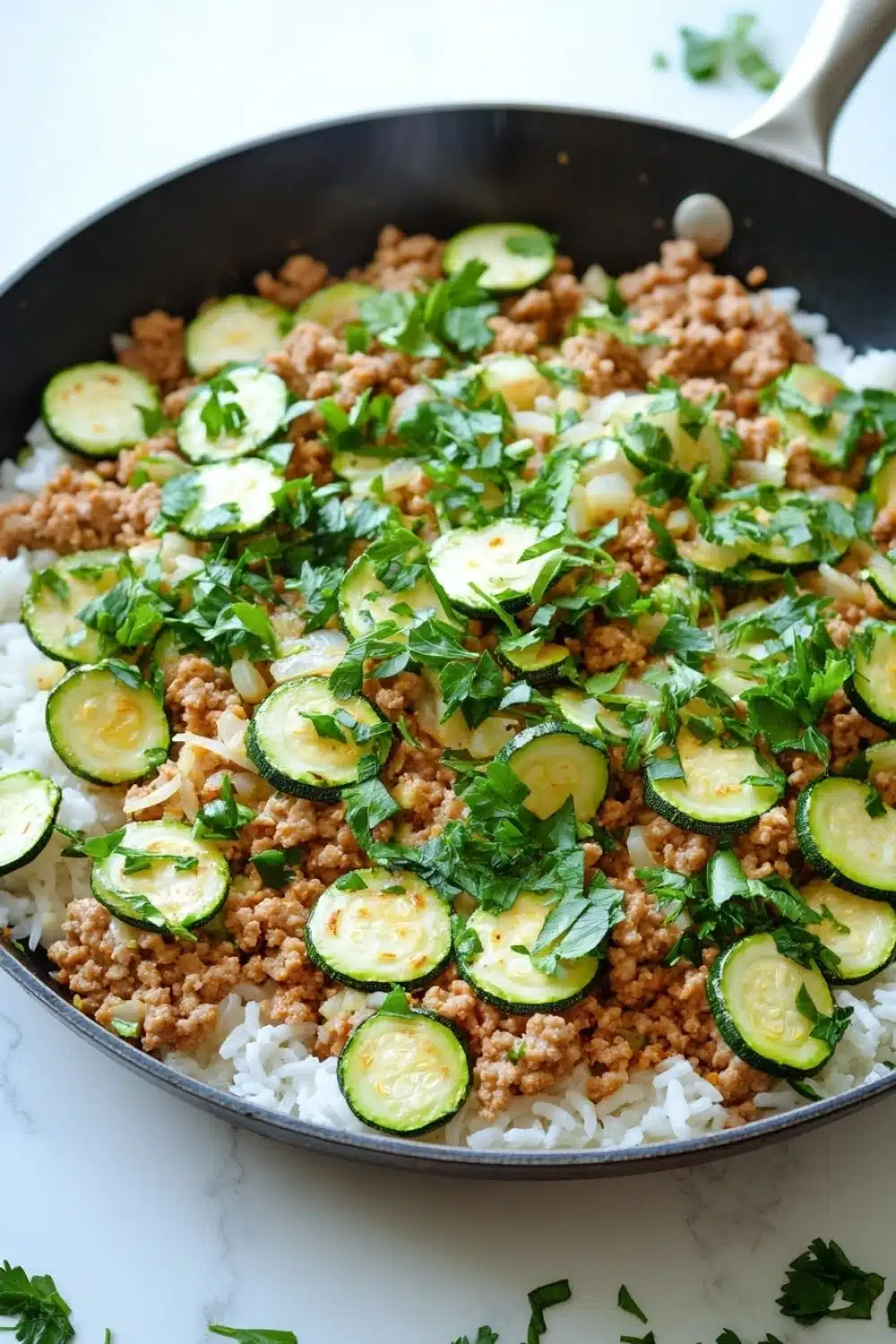 Ground chicken zucchini rice skillet - the image shows a black frying pan with a serving of food in it. the food appears to be a dish made with rice, ground beef, and zucchini. the rice is white and fluffy, and the ground beef is a light brown color. the zucchinis are sliced and arranged in a circular pattern on top of the rice. there are also some chopped parsley scattered around the pan, adding a pop of green color to the dish. the pan is sitting on a white countertop, and there are a few sprigs of parsley on the side.