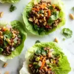 High protein beef lettuce wraps - the image shows four lettuce wraps filled with ground beef and vegetables. the lettuce wraps are arranged on a white marble surface with scattered peanuts and cilantro around them. the ground beef is a dark brown color and appears to be seasoned with herbs and spices. the vegetables include carrots, celery, onions, and peanuts. the wraps are garnished with a sprig of cilantro. the overall appearance of the wraps is fresh and appetizing.