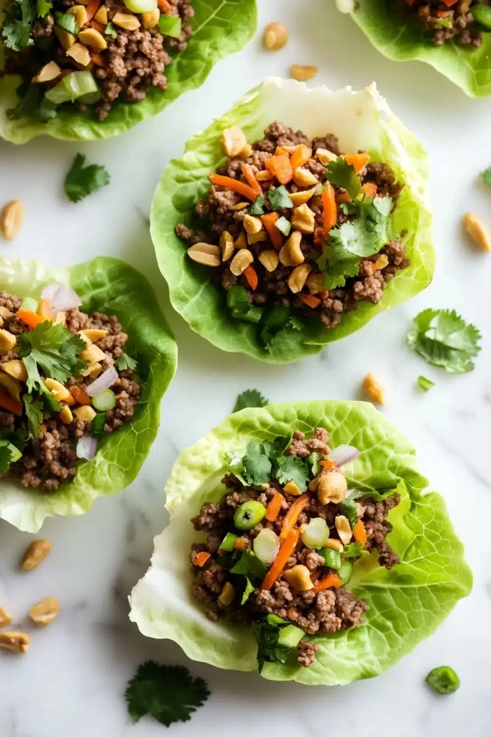 High protein beef lettuce wraps - the image shows four lettuce wraps filled with ground beef and vegetables. the lettuce wraps are arranged on a white marble surface with scattered peanuts and cilantro around them. the ground beef is a dark brown color and appears to be seasoned with herbs and spices. the vegetables include carrots, celery, onions, and peanuts. the wraps are garnished with a sprig of cilantro. the overall appearance of the wraps is fresh and appetizing.