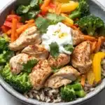 High protein chicken and rice bowl - the image is a close-up of a bowl of food. the bowl is made of gray ceramic and is filled with a colorful mix of rice, vegetables, and chicken. the vegetables include broccoli, carrots, bell peppers, and red bell peppers. the chicken is cooked to a golden brown color and is garnished with a sprig of cilantro. there is also a dollop of sour cream on top of the vegetables. the dish appears to be freshly prepared and ready to eat.