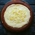 Lemon cake lemon pudding - the image is a close-up of a round cake on a black plate. the cake appears to be freshly baked and has a golden brown crust on top. the top of the cake is covered in a creamy white frosting, which is drizzled with a yellow lemon zest. the frosting is spread evenly over the cake, creating a smooth and even texture. the plate is placed on a dark wooden surface, and the background is blurred, making the cake the focal point of the image.