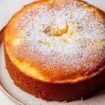Lemon cake with japanese ingredients - the image is of a round cake on a white plate. the cake appears to be freshly baked and has a golden brown crust on top. it is dusted with a generous amount of powdered sugar, giving it a shiny appearance. the plate is sitting on a marble countertop, and the background is blurred, making the cake the focal point of the image.