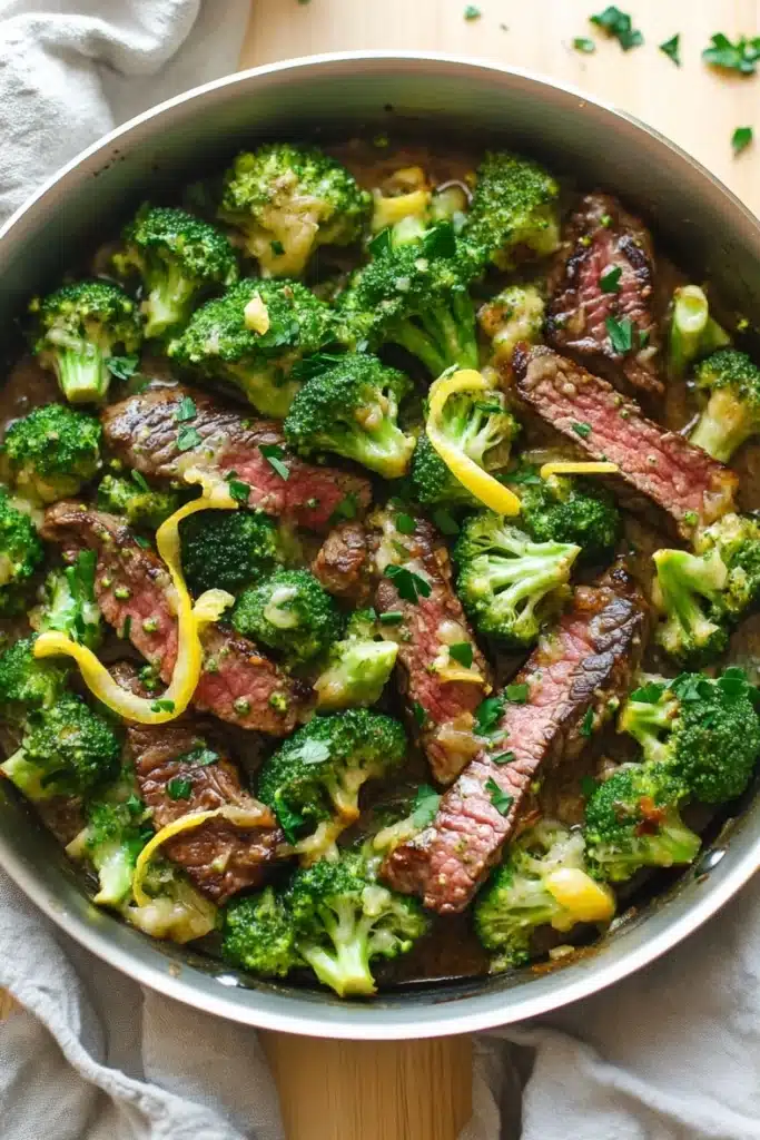 Lemon garlic butter steak and broccoli skillet - the image shows a large stainless steel skillet filled with cooked beef and broccoli. the beef appears to be medium-rare and has a pinkish-brown color. the broccoli is bright green and looks fresh and healthy. the skillet is sitting on a wooden cutting board with a white cloth napkin on the side. there are also some chopped herbs scattered around the skillet. the overall appearance of the dish is savory and appetizing.