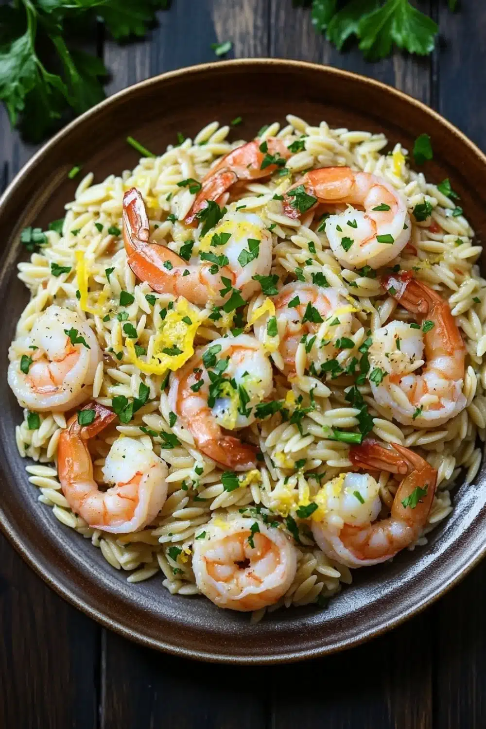 Lemon garlic shrimp orzo skillet - the image is a close-up of a bowl of pasta dish. the pasta appears to be fusilli, which is a type of italian pasta, and is mixed with shrimp. the shrimp are cooked medium-sized and have a pinkish-orange color. they are arranged in a circular pattern on top of the pasta, with some of them overlapping each other. the dish is garnished with chopped parsley and a sprinkle of lemon zest. the bowl is made of dark brown ceramic and is sitting on a wooden table. there are a few sprigs of parsley scattered around the bowl.