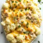 Low carb cheesy tuna casserole - the image is a close-up of a dish of mashed cauliflower. the cauliflower is white and fluffy, and it is topped with melted cheese and chopped parsley. the cheese is a bright yellow color and appears to be melted and bubbly. the dish is served on a white marble countertop. there are a few sprigs of parsley scattered around the dish, adding a pop of green color to the image.