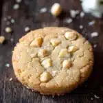 Macadamia nut shortbread cookie - the image is a close-up of a round cookie on a wooden surface. the cookie appears to be freshly baked and has a golden-brown color. it is covered in small white nuts, which are scattered around the edges of the cookie. the nuts are arranged in a criss-cross pattern, with some overlapping each other. the background is blurred, but it seems to be a rustic wooden table with more nuts scattered around. the overall mood of the image is warm and inviting.