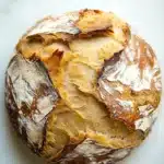 No knead bread new york times - the image is a close-up of a round loaf of bread on a white marble surface. the bread appears to be freshly baked, with a golden-brown crust and a soft, fluffy interior. the edges of the bread are slightly frayed, indicating that it has been baked in the oven. there is a small amount of powdered sugar on top of the crust, giving it a textured appearance. the background is blurred, making the bread the focal point of the image.